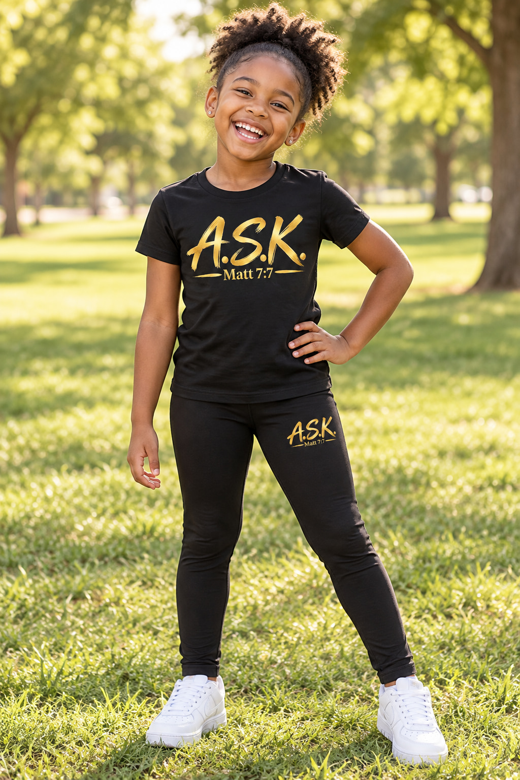 A laughing young Black girl in a sunny park wearing a matching black A.S.K. kids two-piece set — a black tee and black leggings, each printed with the gold A.S.K. Matt 7:7 mark
